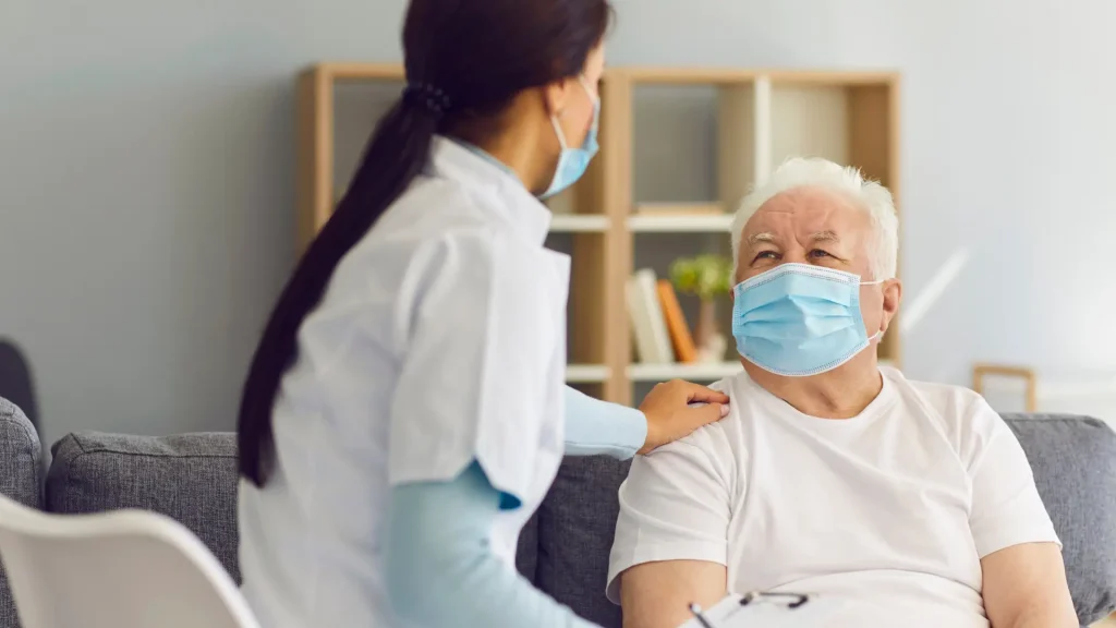 A masked nurse comforting an elderly patient wearing a mask in a home setting.